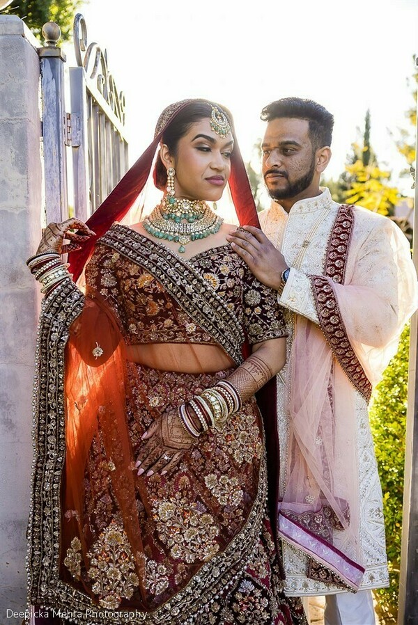 indian couple in red and ivory,bride holding groom,red bridal dupatta,embroidered lehenga photo,cultural wedding look,indian wedding styling,couple pose by drapes,regal bridal portrait,fashion-forward wedding shot,rich wedding textiles