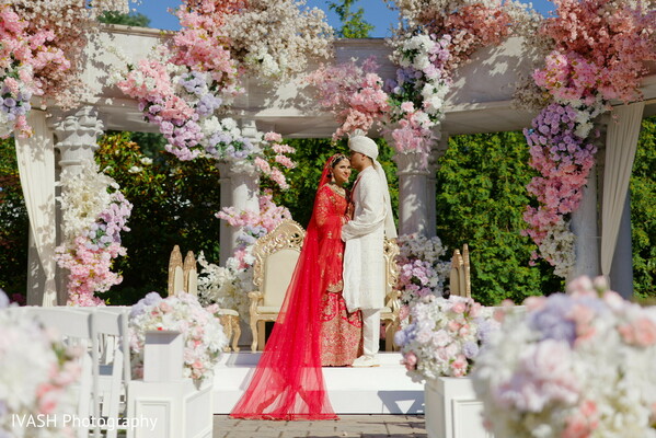 indian bride floral mandap,red bridal lehenga standing,floral arch wedding,pre-ceremony pose,bride at altar entrance,wedding flower canopy,indian outdoor elegance,sacred wedding frame,solo bride moment,mandap entrance portrait