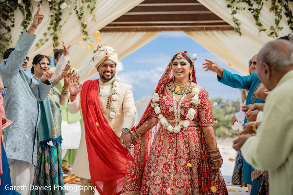 wedding exit joy,indian couple mandap exit,post-ceremony celebration,tropical wedding moment,raised hands bride groom,red lehenga groom celebration,floral canopy marriage,cultural wedding completion,joyful newlyweds photo,indian beach wedding finale