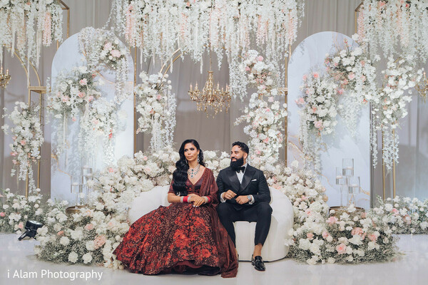 bride groom portrait,floral wedding stage,regal couple shot,red reception gown,wedding throne setup,indian wedding decor,elegant wedding scene,stage with arches,luxury reception backdrop,posed couple moment