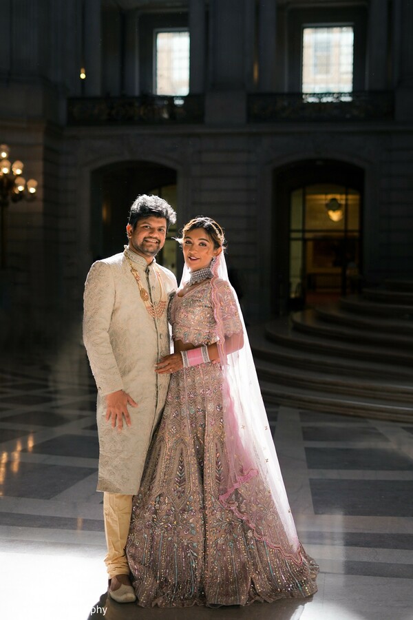 indoor grand venue photo,couple under arch,palace wedding setting,south asian elegance,pink lehenga and sherwani,bride groom pose indoors,elegant architecture wedding,romantic formal shoot,graceful wedding portrait,light-filled couple image