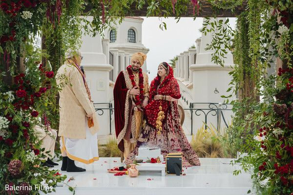 wedding ceremony,indian couple,floral mandap,red bridal lehenga,groom sherwani,joyful wedding moment,garland exchange,south asian rituals,elegant wedding stage,temple backdrop