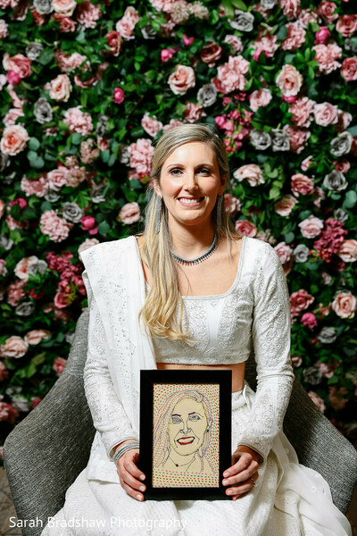 bride with portrait,floral photo wall,custom wedding art,white lehenga bride,bridal portrait,unique wedding details,personalized wedding gift,rose flower backdrop,south asian wedding inspiration,indoor bridal photo