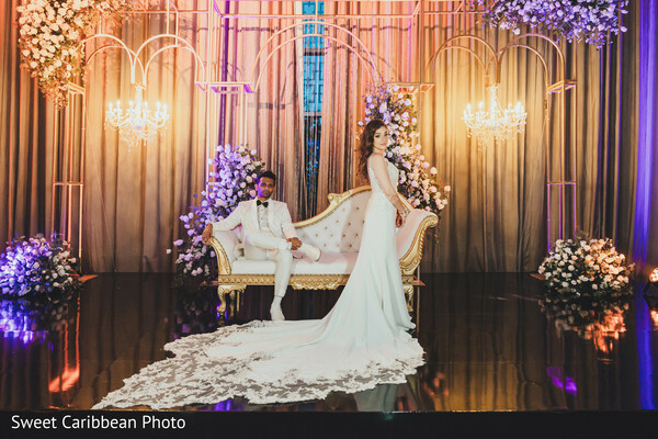 reception stage,bride in white gown,groom in tuxedo,dramatic train,floral backdrop,modern wedding style,romantic lighting,luxury reception,editorial wedding portrait,golden hour glow