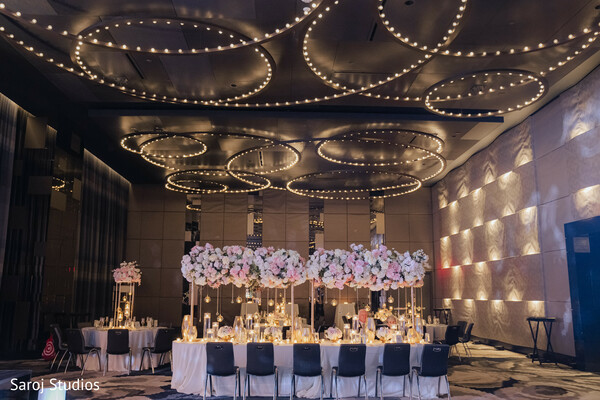 wedding reception decor,luxury banquet table,floral wedding centerpiece,pastel wedding flowers,golden candle decor,indoor wedding reception,modern ceiling lights,romantic wedding setup,luxury wedding venue,elegant wedding tablescape.
