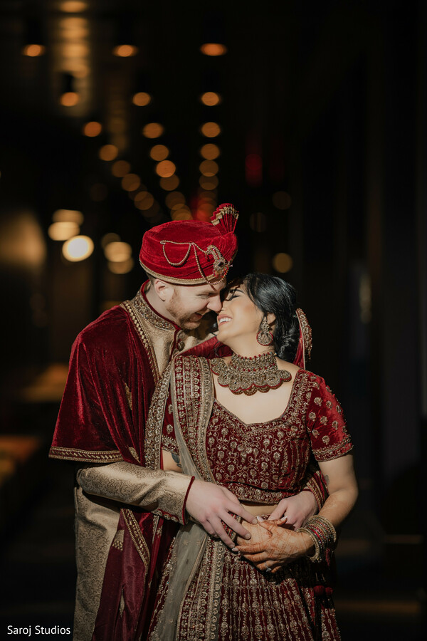 romantic couple embrace,groom holding bride,indian wedding photography,red and gold attire,luxury wedding shot,soft hallway lighting,elegant bridal look,traditional groom attire,romantic wedding photo,timeless wedding love.