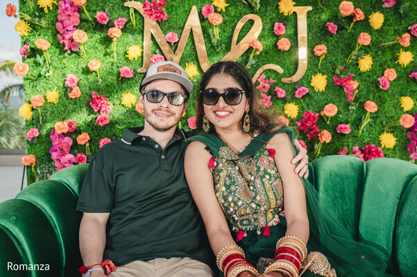 pre-wedding event,couple portrait,floral backdrop,fun wedding moment,indian bride casual look,green and red outfits,wedding signage,relaxed celebration