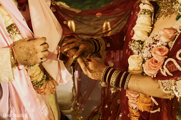 ring ceremony,bride and groom hands,mehndi design,indian jewelry,floral backdrop,intimate moment,cultural wedding,wedding hands,ritual detail,bridal bangles