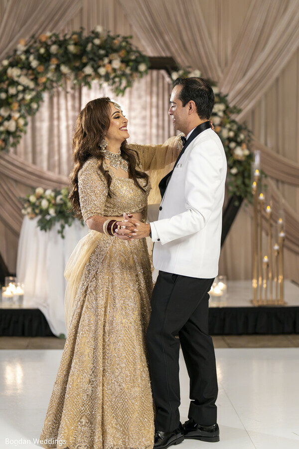 first dance moment,romantic couple portrait,bride in gold lehenga,groom in tuxedo,indoor wedding reception,indian-american wedding,bridal glam,wedding stage backdrop,couple’s first look,elegant wedding moment