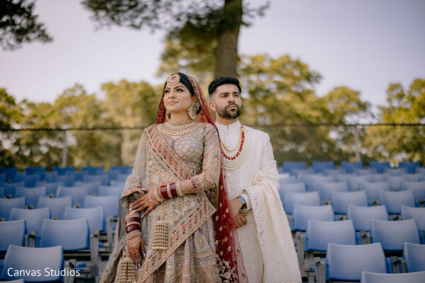 stadium wedding shot,outdoor couple portrait,bridal jewelry,cream and red bridal wear,modern traditional mix,statement photo,bride and groom fashion,power pose,dramatic couple photo,unique location