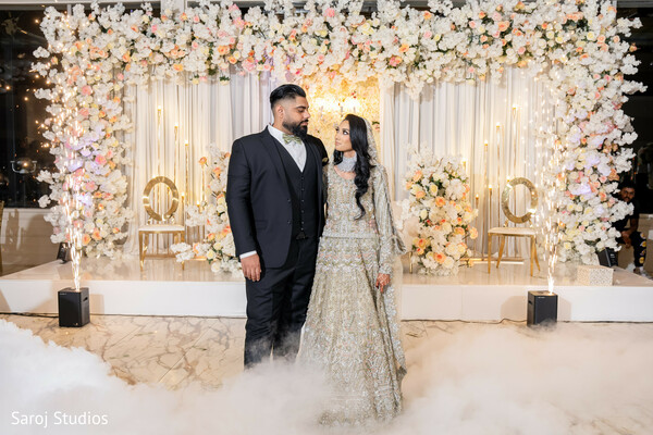 floral wedding backdrop,fog floor effect,couple standing pose,silver bridal gown,black groom suit,luxury wedding setup,elegant wedding stage,romantic wedding portrait,reception decor inspiration,stylish couple photo