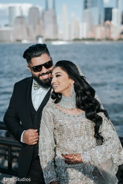 waterfront wedding photo,city skyline backdrop,candid couple laughter,silver gown bride,black suit groom,romantic city photo,joyful wedding moment,stylish urban couple,candid love shot,outdoor wedding setting