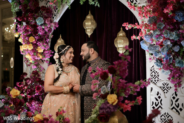 jewel toned wedding decor,floral arch backdrop,bride and groom portrait,pastel bridal outfit,groom in sherwani,romantic floral wedding photo,luxury south asian decor,vibrant wedding photography,colorful wedding stage,couple celebration portrait