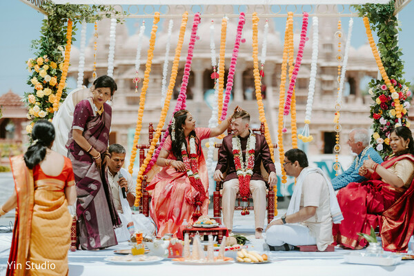 couple standing outside the temple,smiling brightly while holding hands. tags: temple backdrop,newlyweds,couple portrait,wedding joy,holding hands,radiant smiles,indian attire,coral saree,cultural elegance,timeless memory