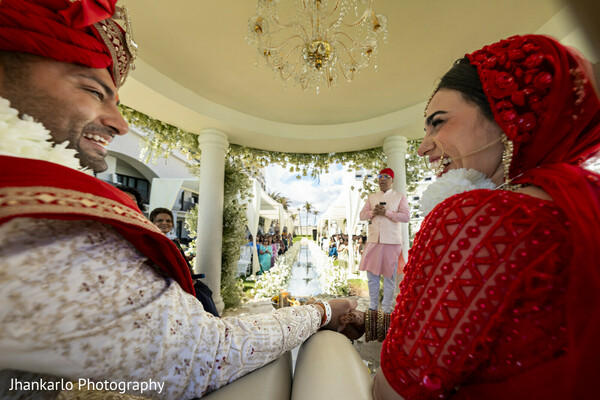 candid laughter,wedding mandap,bride and groom joy,destination wedding,red wedding attire,floral decor,happy couple,cancun wedding,cultural rituals,joyful bride