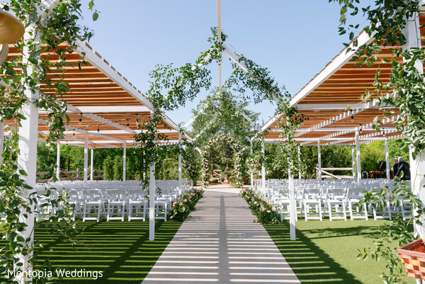 outdoor ceremony,greenery decor,white chairs,modern mandap,aisle view,wedding architecture,natural lighting,open-air venue,symmetrical design,fresh florals