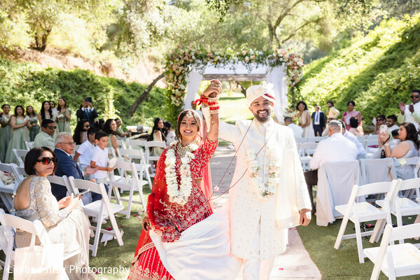 newlyweds,wedding recessional,red bridal lehenga,ivory sherwani,outdoor ceremony,south asian couple,joyful moment,family celebration,floral decor,cultural wedding