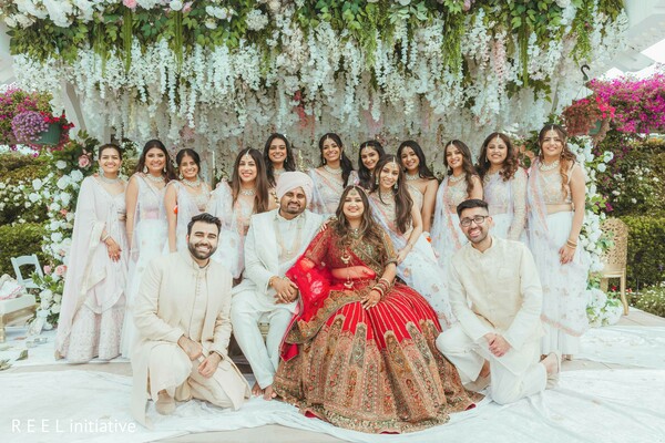 wedding party,coordinated outfits,red bridal lehenga,ivory attire,friends and family,group portrait,south asian wedding,joyful gathering,floral backdrop,candid smiles