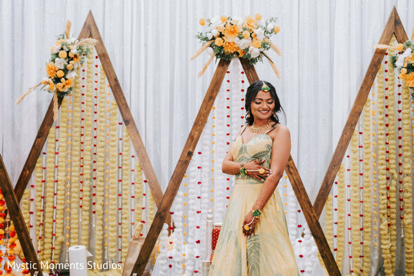 mehndi ceremony outfit,yellow bridal lehenga,floral mehndi backdrop,green bridal jewelry,pre-wedding portrait,south asian bride styling,marigold string decor,cultural wedding celebration,bride solo portrait,traditional mehndi look