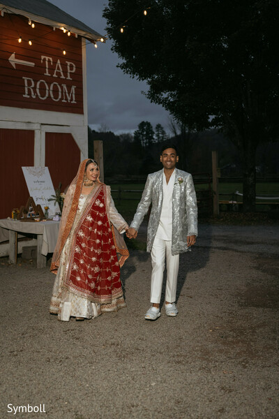 evening wedding portrait,red bridal lehenga,silver groom jacket,rustic tap room venue,string lights ambiance,south asian couple walk,romantic dusk moment,fusion wedding fashion,countryside reception,newlywed exit