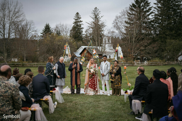 outdoor hindu ceremony,red bridal lehenga,ivory groom sherwani,countryside wedding venue,floral ceremony arch,south asian wedding ritual,seated wedding guests,rustic barn backdrop,multicultural ceremony,sacred vows