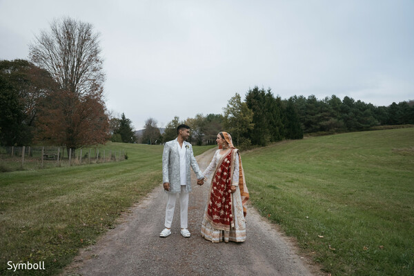 countryside wedding portrait,red dupatta contrast,ivory bridal lehenga,modern silver groom jacket,hand-in-hand couple,south asian fusion wedding,rural landscape backdrop,romantic outdoor moment,newlywed stroll,autumn field scenery