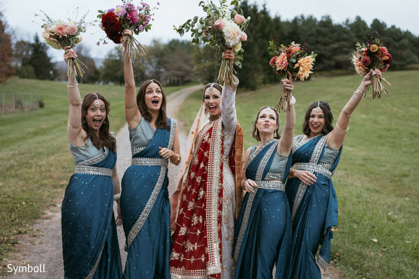 bridesmaids in blue saris,red bridal lehenga,outdoor countryside wedding,bouquet toss moment,bridal squad celebration,south asian bridal party,joyful wedding energy,floral bouquets,wedding sisterhood,rustic landscape backdrop