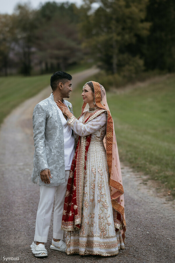 ivory and red bridal lehenga,silver textured sherwani,romantic countryside portrait,intimate eye contact,south asian bride and groom,autumn wedding setting,gravel road backdrop,embroidered dupatta details,cultural wedding attire,soft outdoor lighting