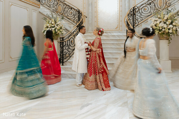 grand staircase wedding,ivory sherwani groom,red bridal lehenga,bridesmaids in motion,luxury venue backdrop,hindu wedding attire,celebratory swirl,marble ballroom setting,cultural wedding fashion,romantic couple portrait
