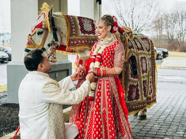 baraat horse,groom kneeling proposal pose,red bridal lehenga,ivory sherwani,ornate horse embroidery,bridal chooda bangles,outdoor wedding entrance,romantic hand clasp,south asian wedding tradition,luxury wedding portrait