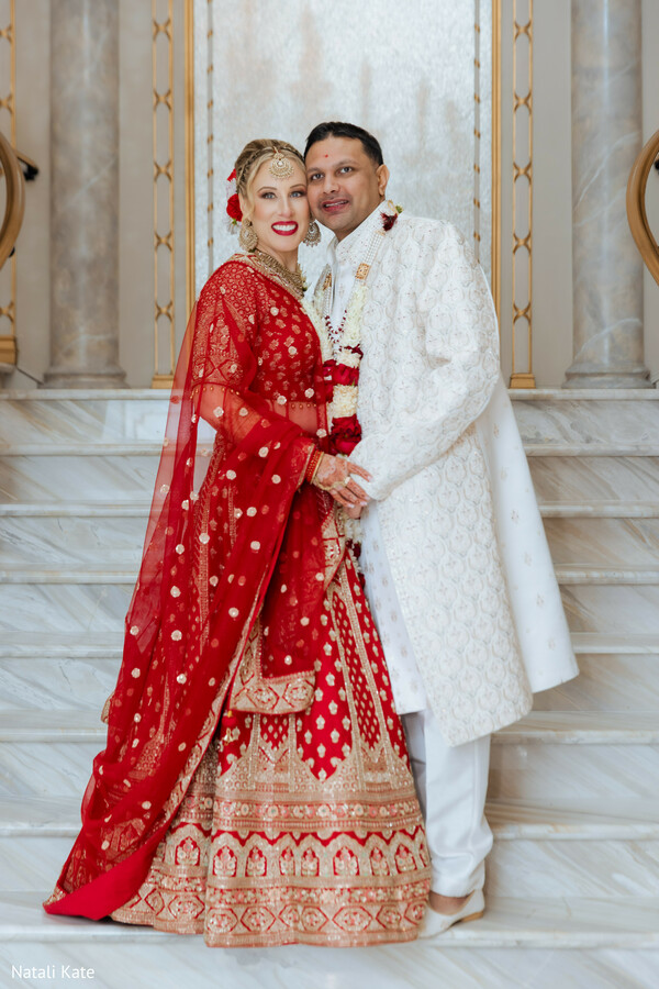 staircase wedding portrait,red and ivory wedding attire,marble venue interior,bridal lehenga detail,sherwani embroidery,luxury hindu wedding,romantic couple pose,gold venue accents,newlywed portrait,elegant indoor ceremony