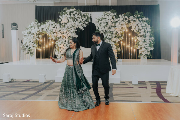 reception couple walk,emerald lehenga detail,black tuxedo elegance,ballroom wedding reception,modern south asian style,coordinated evening attire,floral stage background,confident couple pose,wedding dance moment,luxury reception fashion