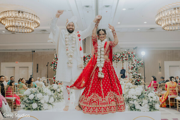 wedding stage celebration,red lehenga twirl,ivory sherwani groom,raised hands victory pose,floral reception backdrop,hindu wedding joy,luxury ballroom chandelier,newlywed celebration moment,garland exchange complete,grand wedding entrance