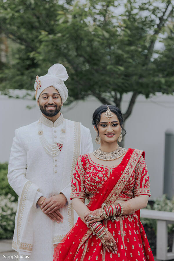 red bridal lehenga,ivory sherwani groom,traditional safa turban,layered pearl mala,bridal chooda bangles,gold maang tikka,outdoor wedding portrait,embroidered zari work,hindu wedding attire,elegant couple pose