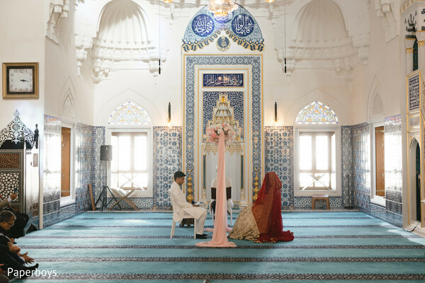 nikah ceremony,mosque wedding setting,red bridal lehenga,ivory sherwani groom,islamic wedding ritual,blue tile architecture,bridal dupatta over head,sacred marriage moment,traditional muslim wedding,cultural ceremony setting