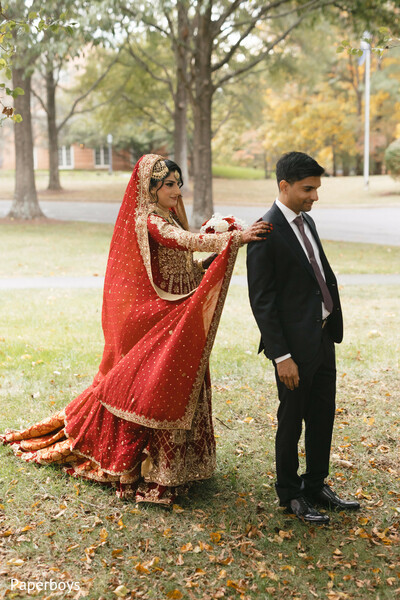 first look wedding moment,playful bridal approach,flowing red bridal train,autumn outdoor setting,groom anticipation pose,traditional bridal attire,romantic wedding reveal,south asian bride portrait,intimate pre-ceremony moment,fall wedding photography