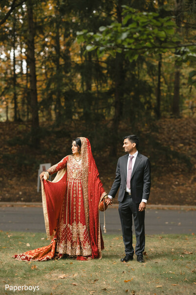 red bridal lehenga,autumn wedding backdrop,flowing bridal dupatta,outdoor couple portrait,gold embroidery detail,modern groom suit,fall foliage setting,traditional south asian bride,romantic hand hold,bridal train detail