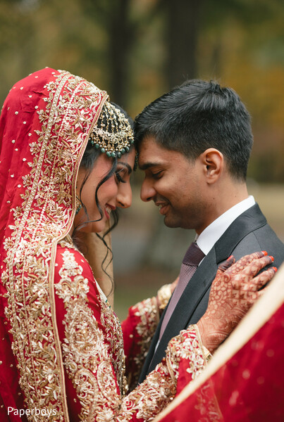 forehead touch moment,bridal mehndi hands,gold embroidered dupatta,intimate couple portrait,red bridal attire,emotional wedding moment,traditional bridal jewelry,outdoor fall romance,close-up wedding photography,south asian bridal elegance