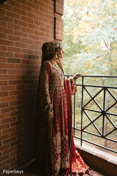 balcony bridal portrait,red and gold lehenga,autumn wedding colors,dramatic bridal dupatta,ornate bridal jewelry,south asian bridal fashion,outdoor elegance,flowing bridal train,fall foliage background,regal bridal pose