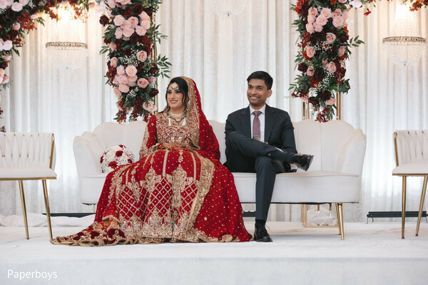 reception stage seating,red bridal lehenga,groom in dark suit,floral stage backdrop,modern south asian reception,wedding sofa portrait,blush and burgundy florals,cultural wedding elegance,bride and groom pose,contemporary wedding style