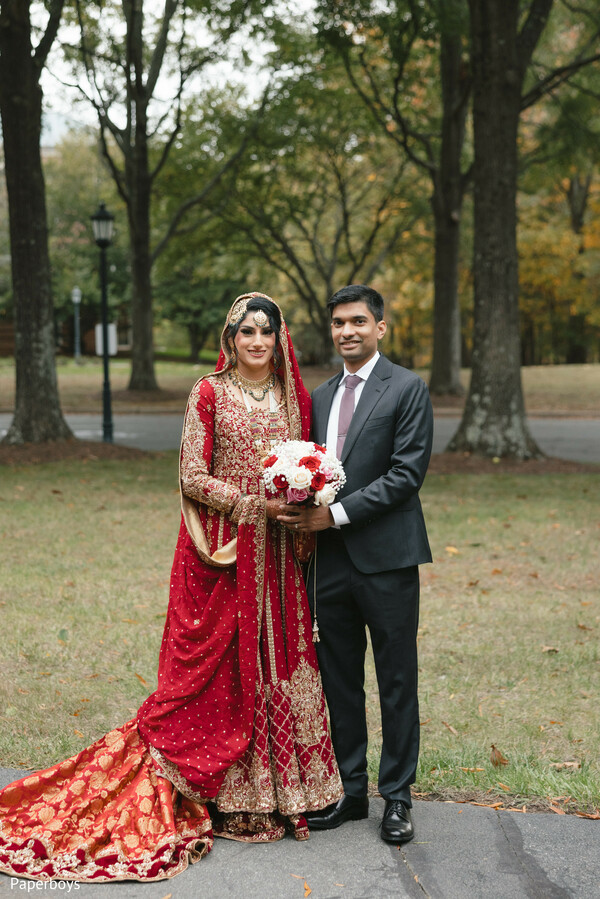 outdoor wedding portrait,red bridal train,rose bouquet detail,fall wedding setting,traditional south asian bride,modern groom suit,romantic couple pose,embroidered lehenga,cultural bridal elegance,autumn ceremony vibe