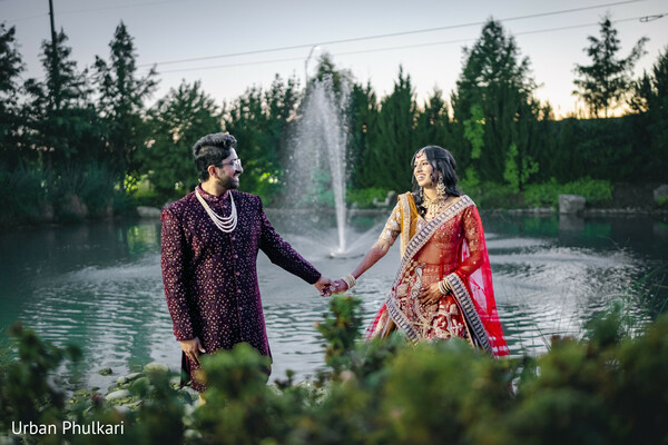 lakeside couple portrait,maroon sherwani groom,red and gold bridal lehenga,pearl mala necklace,romantic hand hold,sunset engagement session,outdoor wedding photography,flowing bridal dupatta,fountain backdrop,south asian pre-wedding shoot