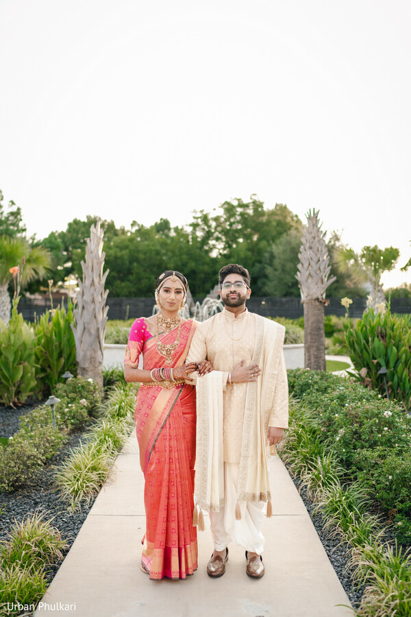 pink bridal saree,cream kurta groom,outdoor garden portrait,traditional south asian attire,gold bridal jewelry,couple walkway pose,pre-wedding photoshoot,elegant saree drape,tropical landscaping backdrop,coordinated wedding fashion