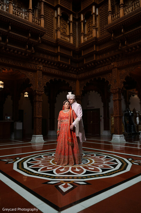 heritage wedding architecture,ornate palace interior,orange bridal lehenga,ivory sherwani groom,cultural wedding portrait,symmetrical wedding photography,luxury indian wedding venue,temple architecture backdrop,elegant couple portrait,traditional wedding setting
