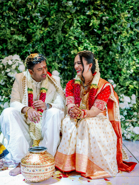 hindu wedding ritual,couple laughing ceremony,floral backdrop greenery,kalash ritual,bride saree red white,groom sherwani cream,candid wedding moment,indian ceremony traditions,joyful couple,wedding rituals