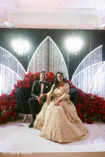 wedding stage couple seated,red floral decor,reception throne couch,luxury wedding backdrop,bride gold lehenga,groom tuxedo,glamorous wedding setup,elegant reception portrait,floral stage design,couple seated