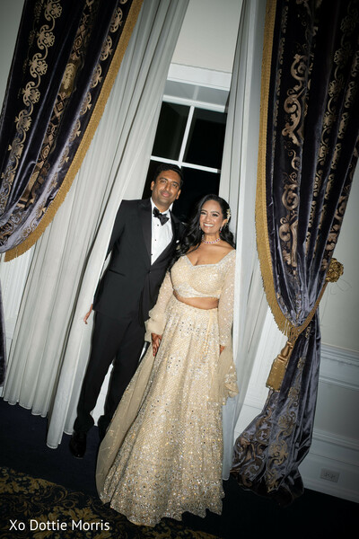 couple portrait indoor,reception outfit bride lehenga,groom tuxedo black,elegant drapery backdrop,formal wedding portrait,evening wedding look,indian bride gold lehenga,groom formal wear,classic wedding style,luxury venue