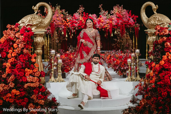 wedding stage portrait,bride red lehenga,groom ivory sherwani,luxury mandap decor,red floral backdrop,regal couple pose,indian wedding fashion,ornate stage design,royal wedding aesthetic,couple portrait