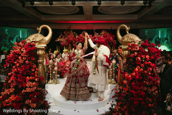 hindu wedding ceremony stage,varmala moment,couple exchanging garlands,red floral decor,gold swan statues,indian wedding rituals,joyful ceremony moment,luxury mandap design,wedding celebration,cultural traditions