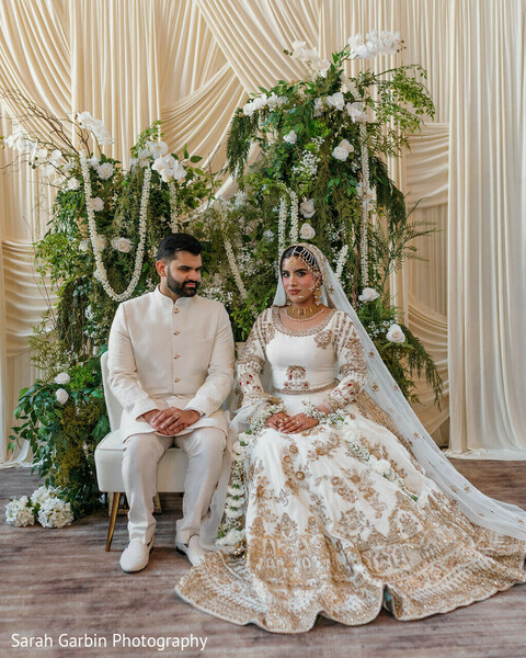 greenery backdrop wedding,couple seated portrait,ivory gold lehenga,groom ivory sherwani,floral greenery decor,romantic indoor setup,elegant wedding styling,neutral tones wedding,soft floral installation,indian wedding portrait